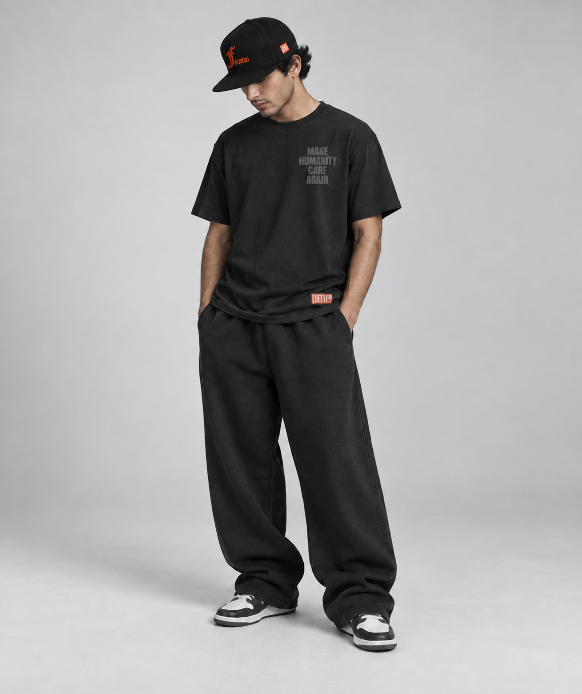A man modeling a black crewneck t-shirt, black sweatpants, a black cap, and black and white sneakers against a light gray studio background.