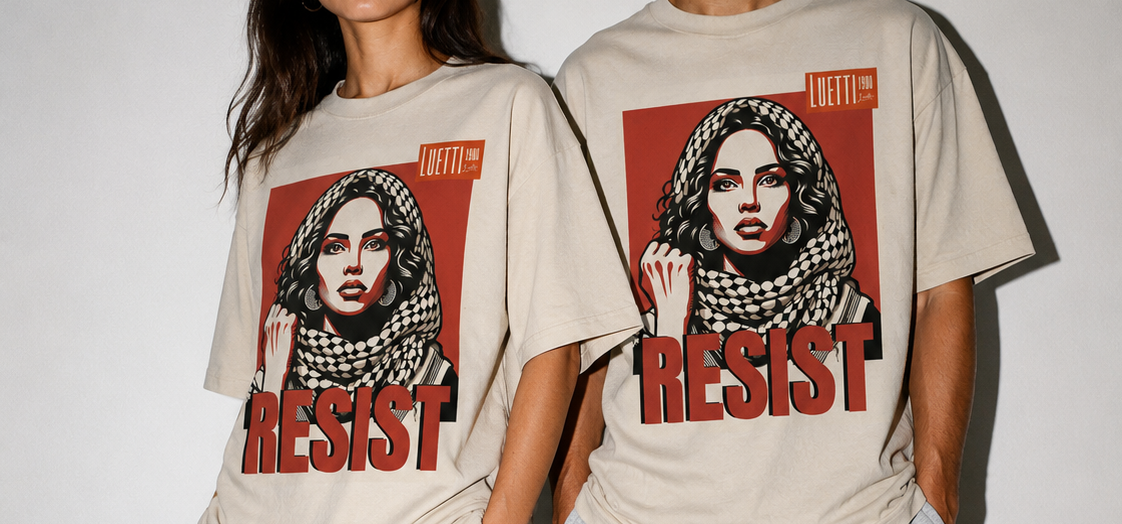 Two models wearing matching beige graphic t-shirts with a red and black print, styled with white sneakers and either white or gray sweatpants against a plain white wall.