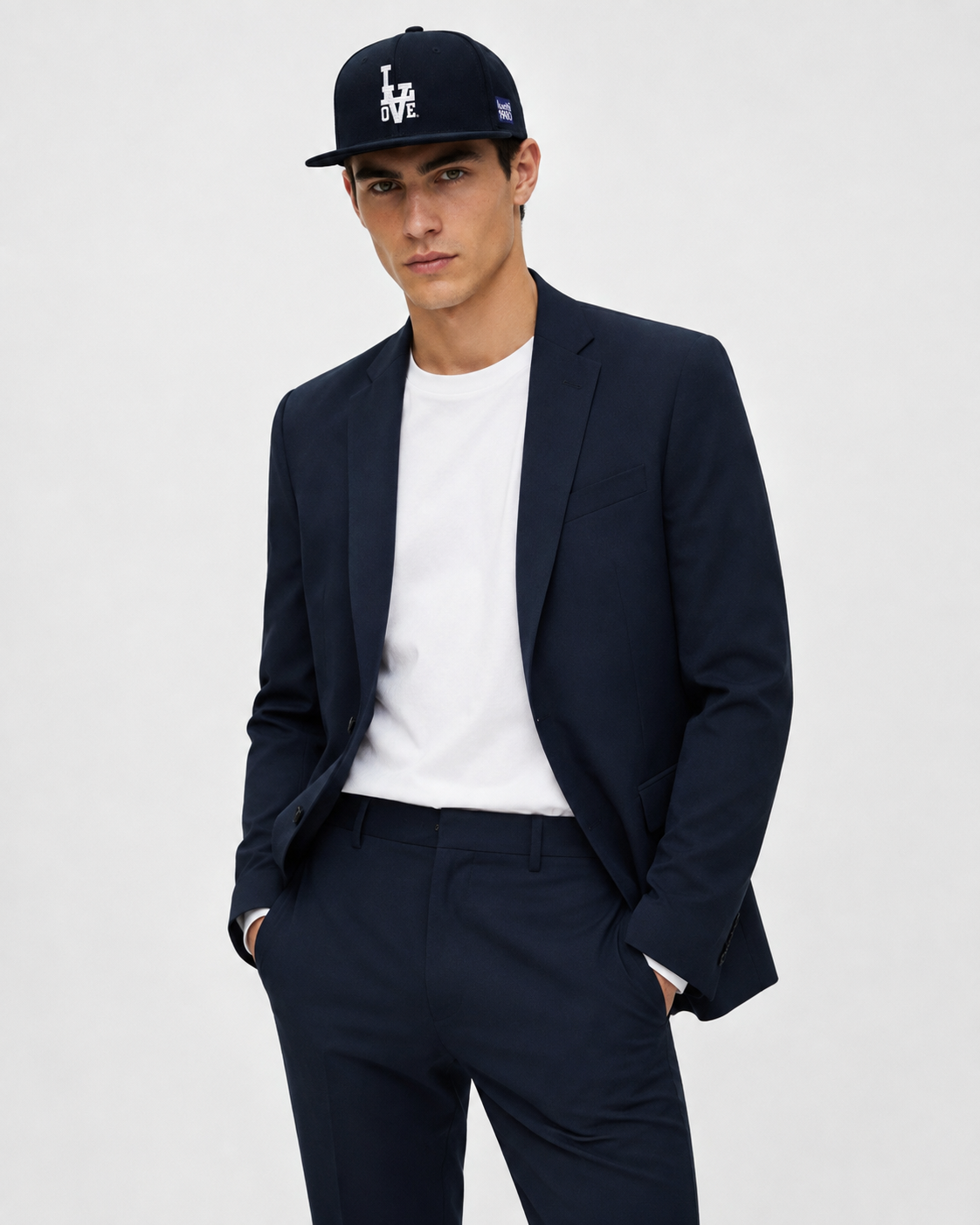 A man wearing a navy blue suit jacket and matching trousers, styled with a white t-shirt and a navy blue baseball cap with a white 'LOVE' logo, set against a plain light gray background.