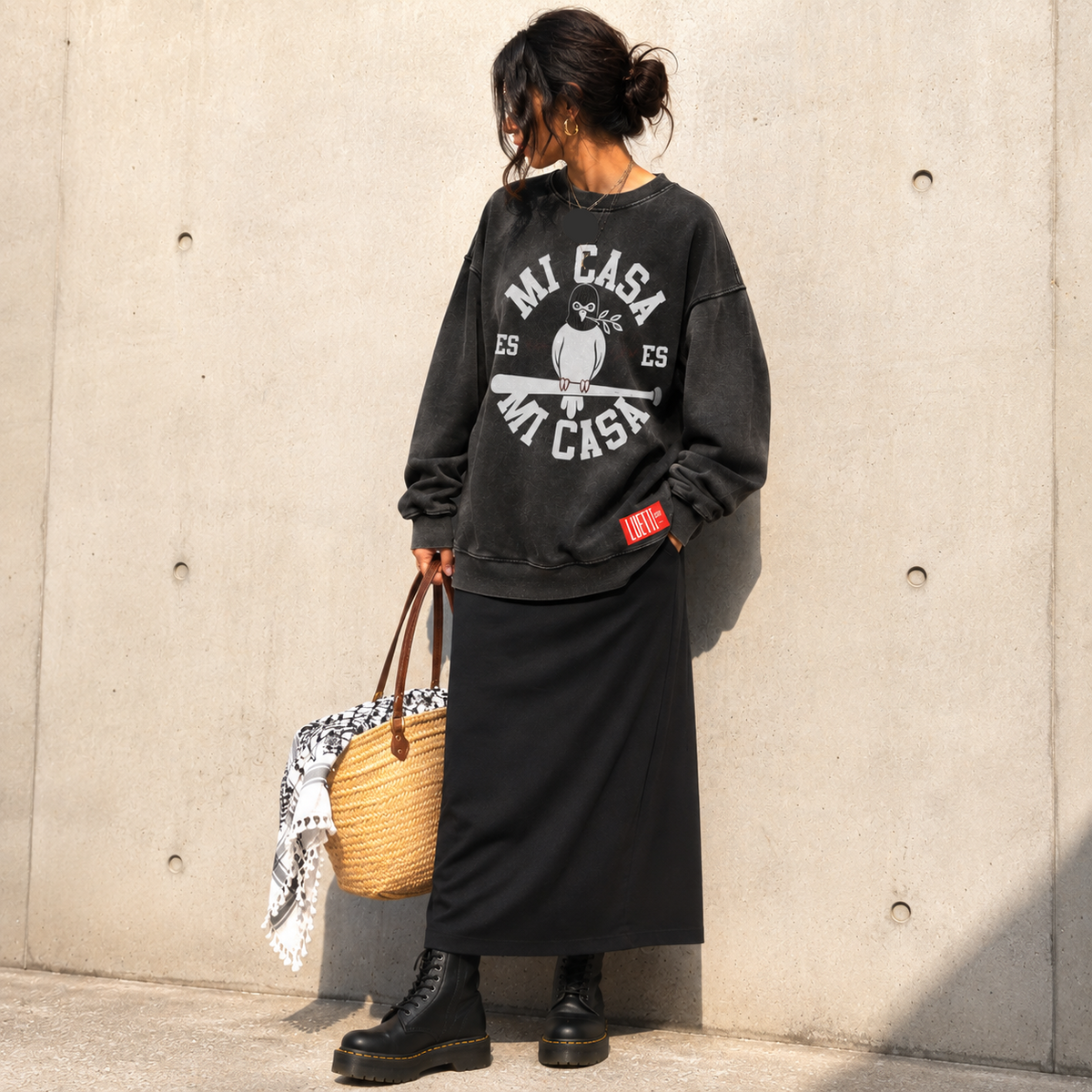 A woman stands against a concrete wall wearing an oversized black sweatshirt with a distressed white graphic that reads 'Mi Casa Es Mi Cisa' and a small red 'FATE' patch on the sleeve, paired with a long black skirt and black boots. She is holding a woven straw tote bag with a black-and-white patterned scarf draped over it.