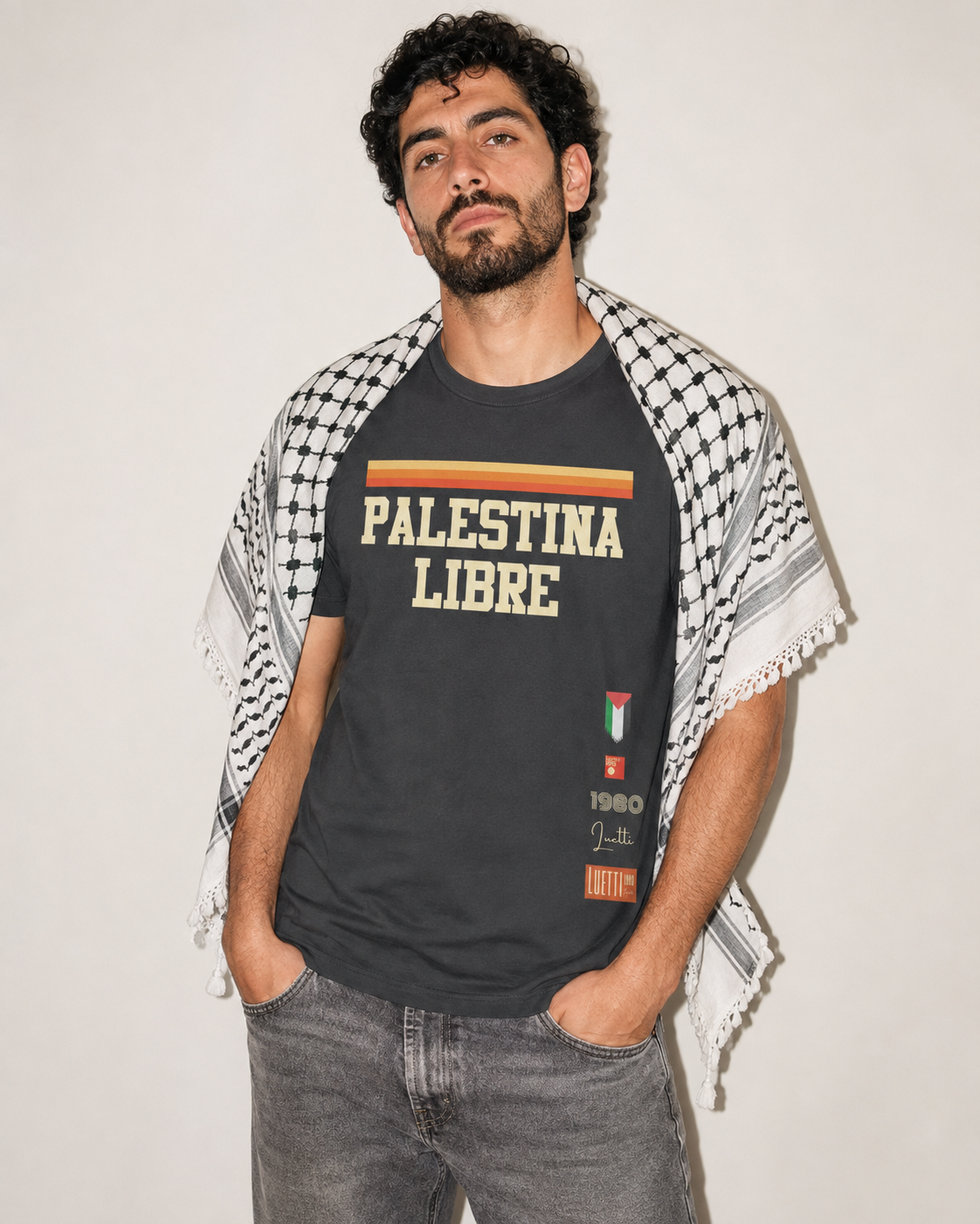 A man wearing a black graphic t-shirt with 'PALESTINA LIBRE' printed in beige and orange, styled with a black-and-white keffiyeh shawl draped over his shoulders and gray jeans, set against a plain off-white background.