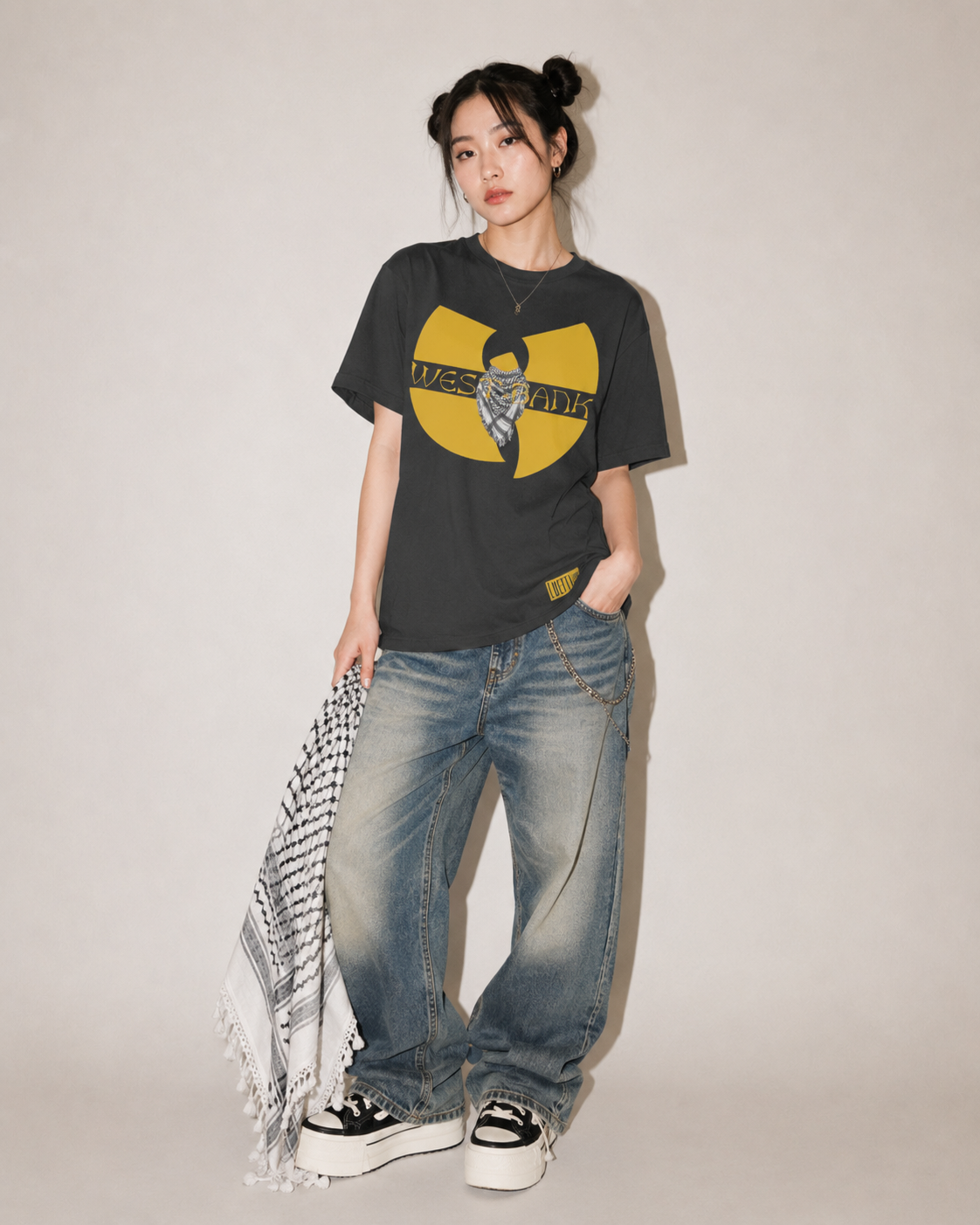 A model stands against a plain light beige studio background wearing a black graphic t-shirt with a yellow and black Wu-Tang Clan logo, paired with wide-leg blue jeans, black and white platform sneakers, and a black-and-white patterned scarf.
